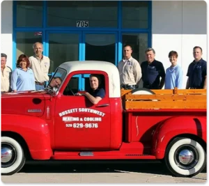 A group of eight people pose in front of a building with blue doors, with a red Russett Southwest Heating & Cooling truck parked in front.